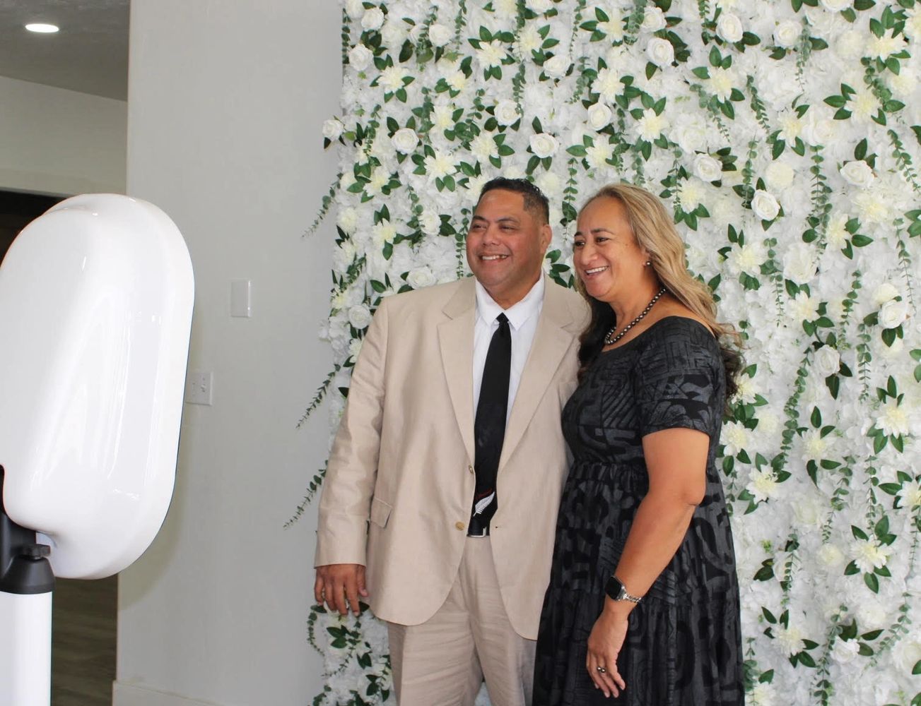 A smiling couple poses in front of a white flower wall backdrop.