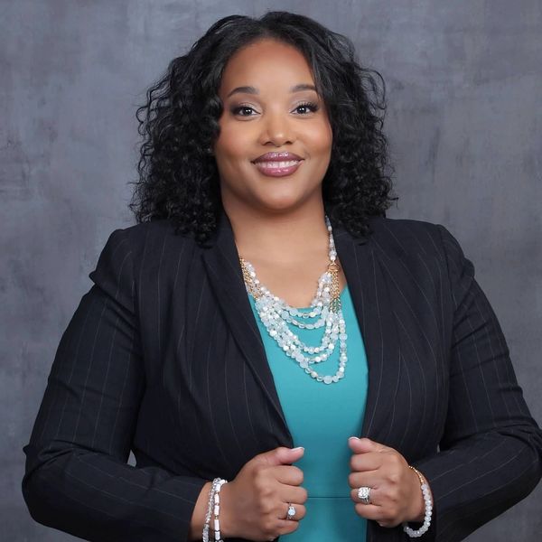 Confident woman in teal dress and black blazer posing with jewelry.