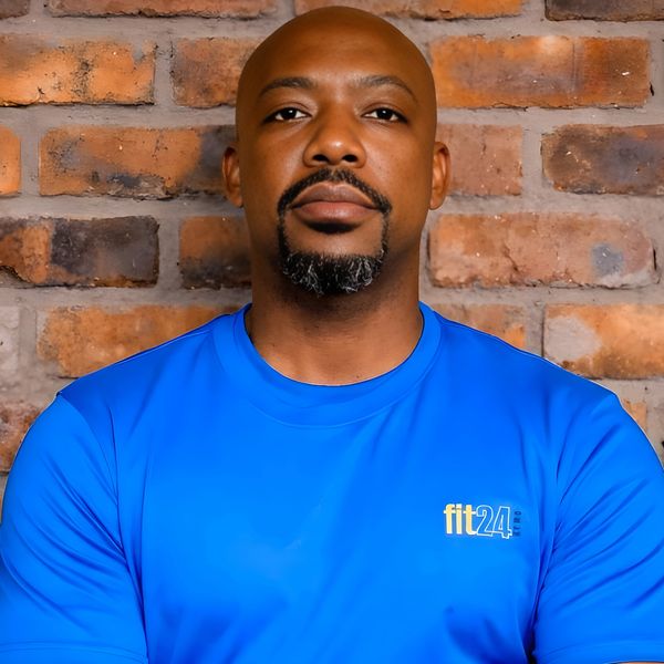 Confident man in a blue fit24 t-shirt standing against a brick wall.