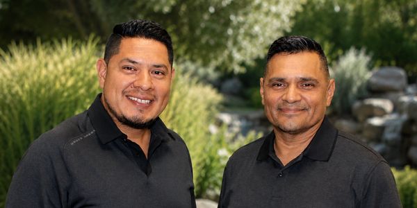 Two smiling men in black polo shirts outdoors with greenery in the background.