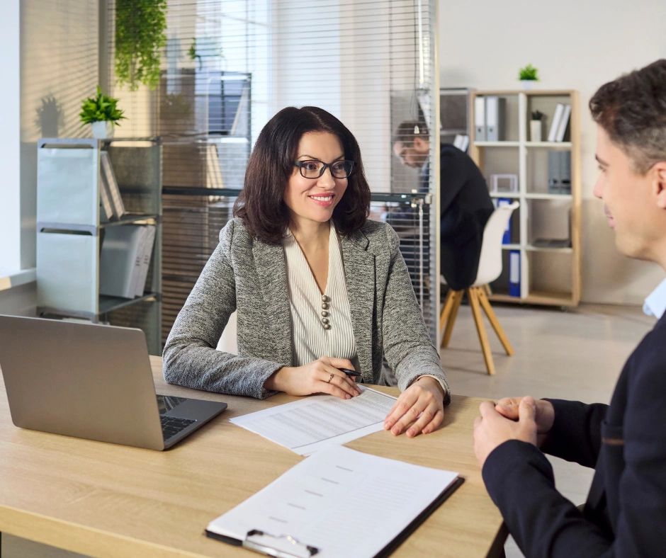 A woman in an office interviewing a man across the table with documents and laptop.