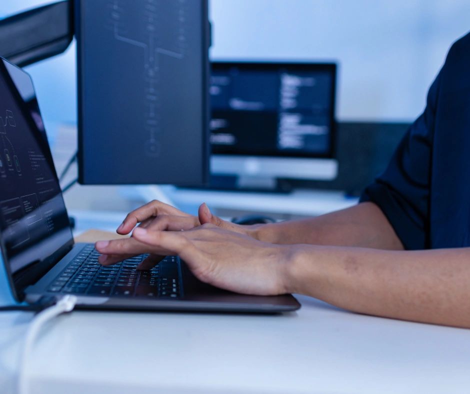 Person typing code on a laptop with multiple monitors in the background.