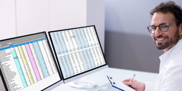 Man analyzing data on two monitors with spreadsheets, smiling.