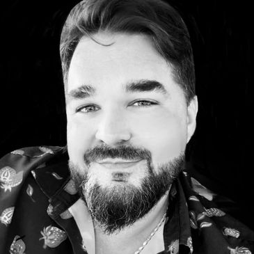 Portrait of a smiling man with a beard and chain necklace, wearing a strawberry-patterned shirt.