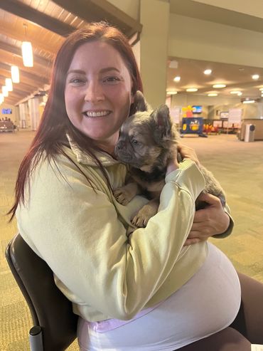 A smiling woman in a yellow sweater holds a small, fluffy puppy indoors.