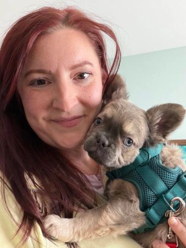 Woman with red hair holding a small dog in a teal harness.