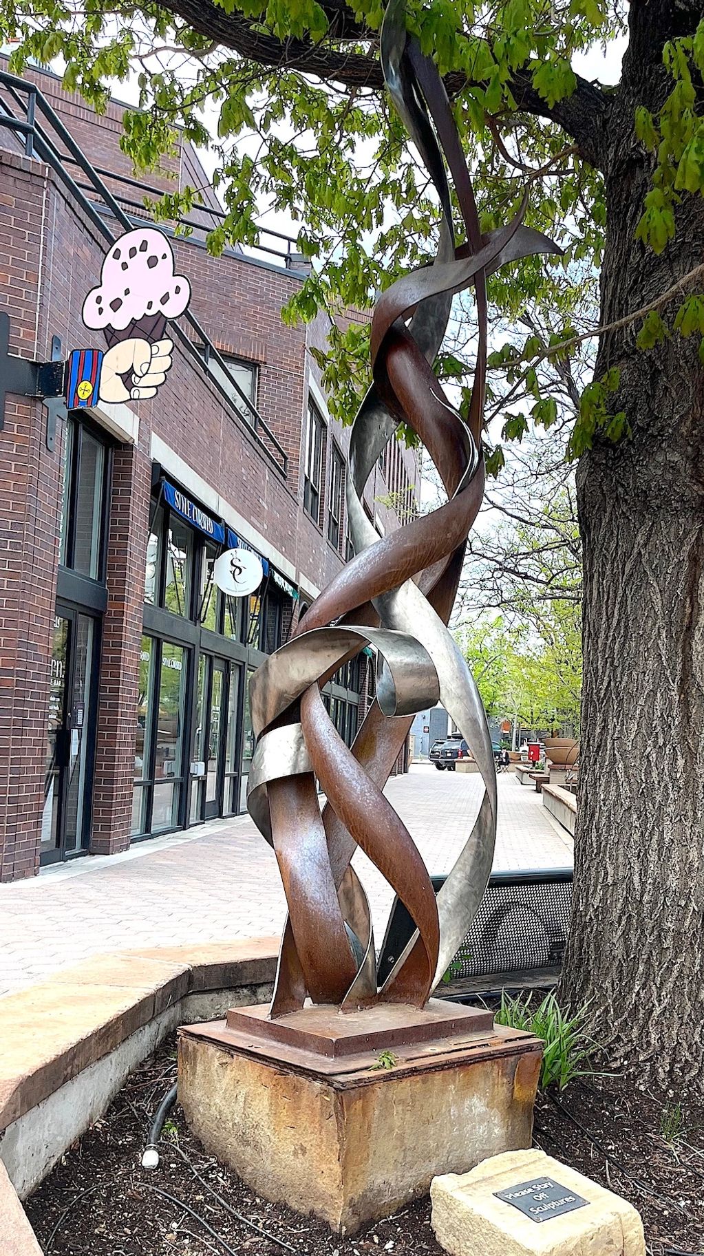 Abstract twisted metal sculpture in an urban park setting beside a tree.