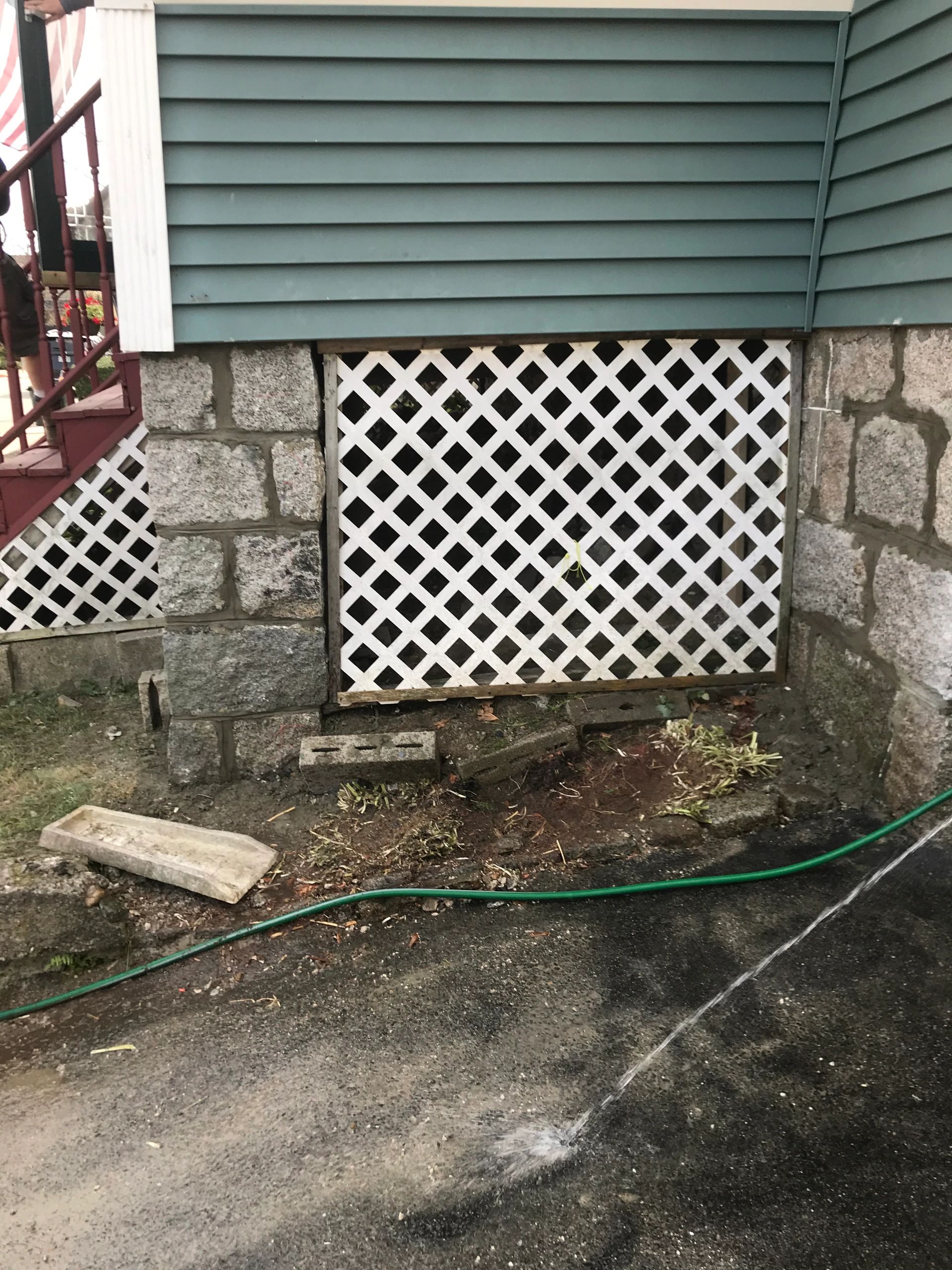 Stone wall with white lattice and green siding beside a garden hose spraying water.