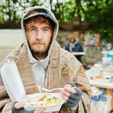 A homeless man wrapped in a blanket holding a food container outdoors.