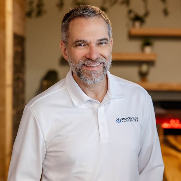 Smiling man in a white Victoria Fide Consulting polo shirt indoors.