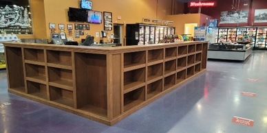 Empty wooden shelves in a store aisle with refrigerated sections in the background.