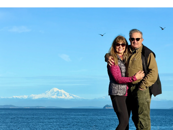 Couple embracing with ocean and snow-capped mountain in background.