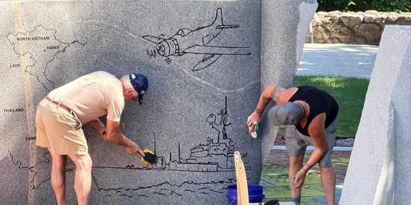 Two men cleaning a war memorial with engraved images.