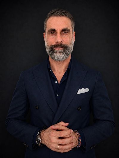 Confident middle-aged man in navy blazer with gray beard against black background.