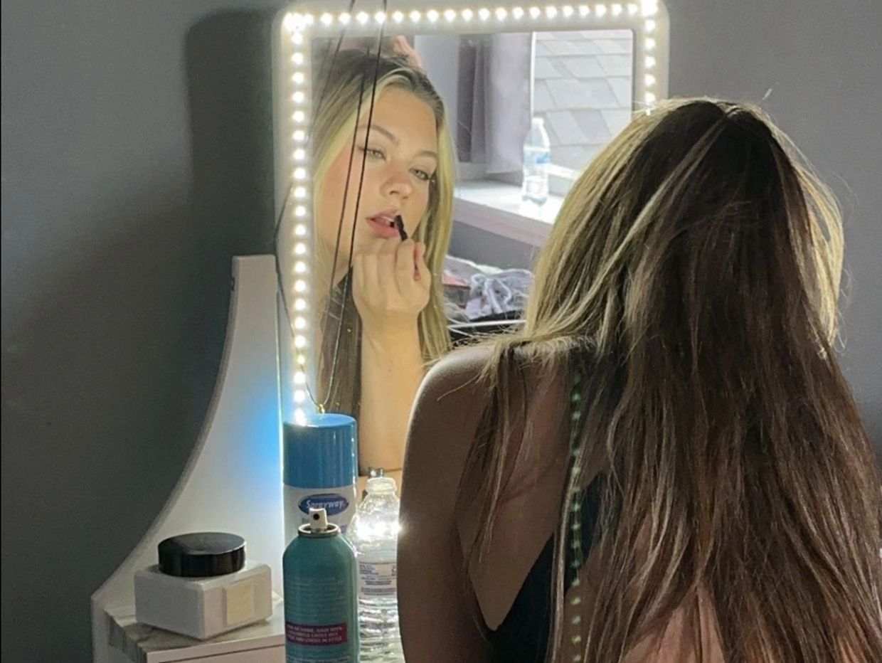 A young woman applying makeup in front of a lighted mirror at a vanity table.