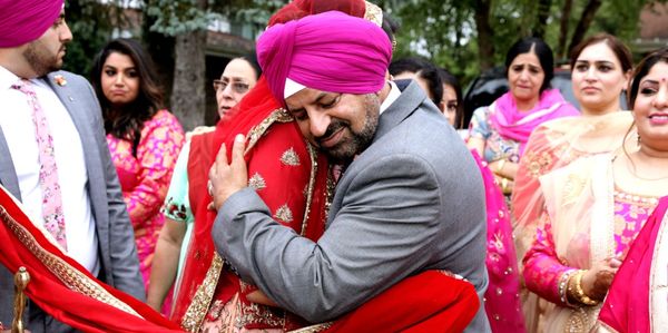 A man in a pink turban warmly embraces a woman in traditional red attire at a festive gathering.
