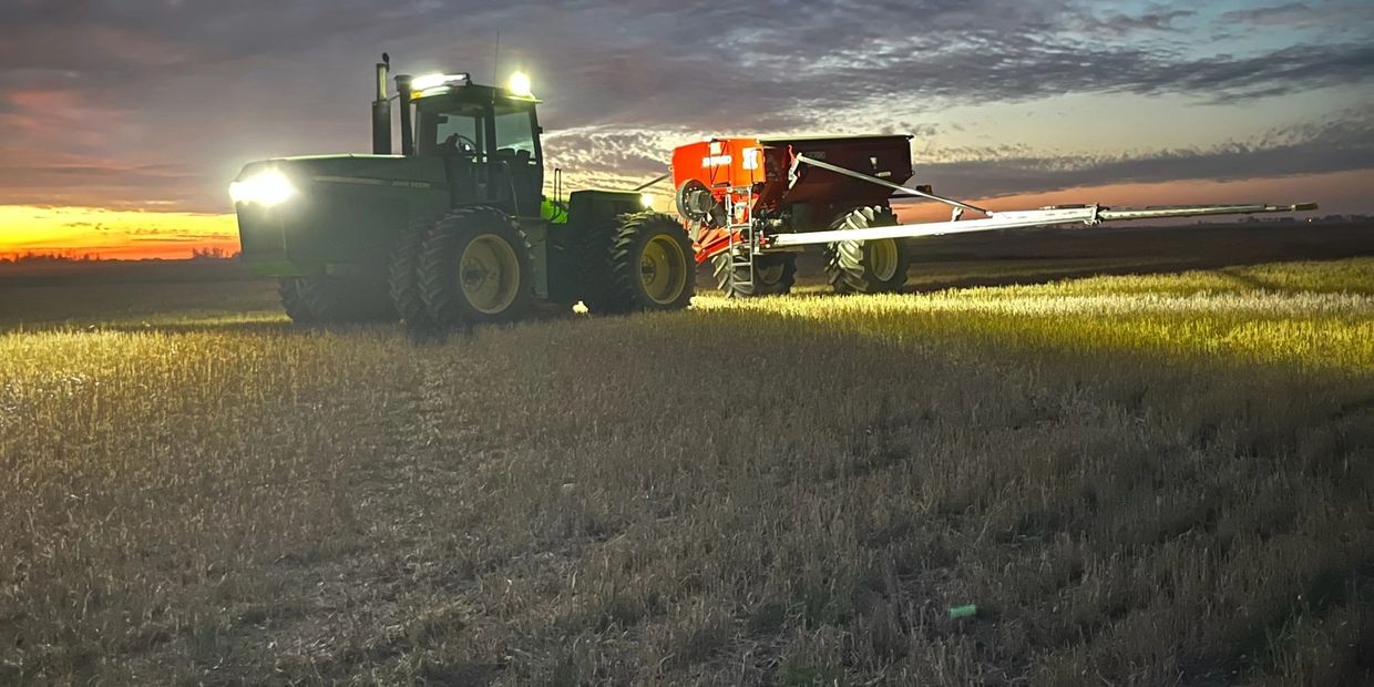 A tractor and red farming equipment work in a field at dusk under a dramatic sky.