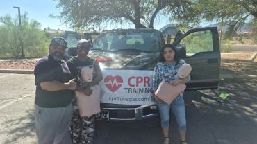 Three people posing with CPR mannequins in front of a vehicle.