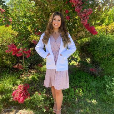 Young woman in a white coat smiles in a garden with blooming pink flowers.