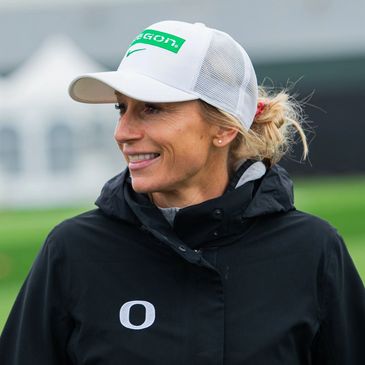 Woman in a white cap and black jacket smiling outdoors.