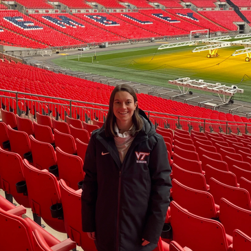 A woman in a VT Soccer jacket stands in a mostly empty red-seated stadium.