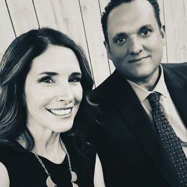Smiling couple dressed formally, posing for a selfie indoors.