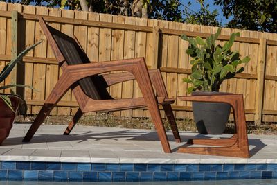 Modern wooden lounge chair and matching stool by a pool with potted plants and wooden fence.