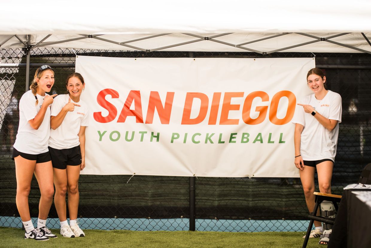 Three girls in white shirts point excitedly at a San Diego Youth Pickleball banner.