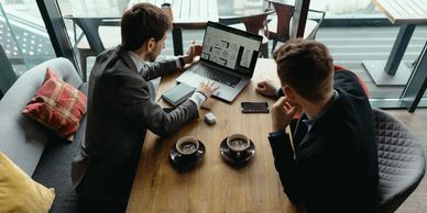 Two men in business attire discussing data on a laptop in a cafe.