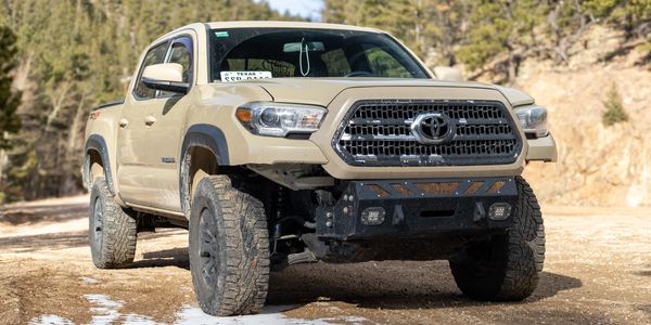 Beige Toyota Tacoma truck on a dirt road with snow patches in a forested area.