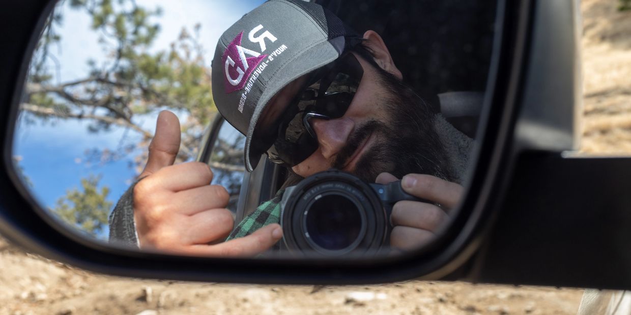 Man with a beard and cap takes a selfie in a car side mirror, giving a thumbs-up.