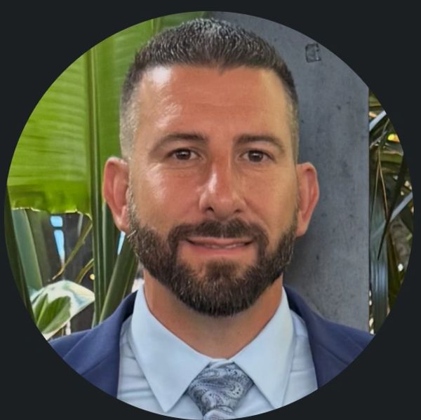 Man with a beard in a suit smiling against a leafy background.