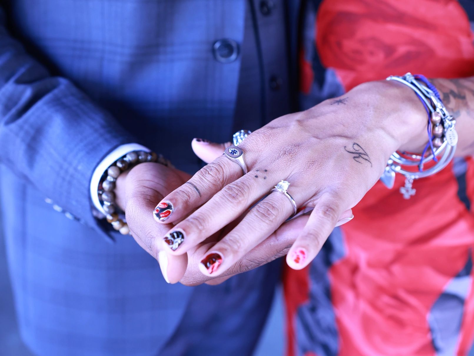 Hands adorned with rings, bracelets, and nail art against a blue suit and red dress background.