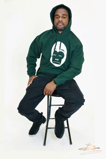 Man in green hoodie with a gorilla face design sitting on a stool.