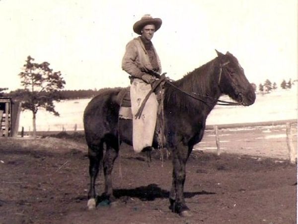 Man in cowboy attire sitting on a horse in a rustic outdoor setting.
