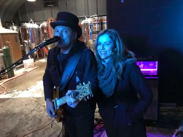 A man playing guitar and singing with a woman smiling beside him.