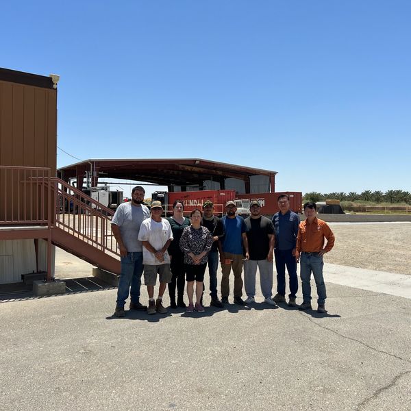 Group of nine people standing outside near industrial buildings on a clear day.