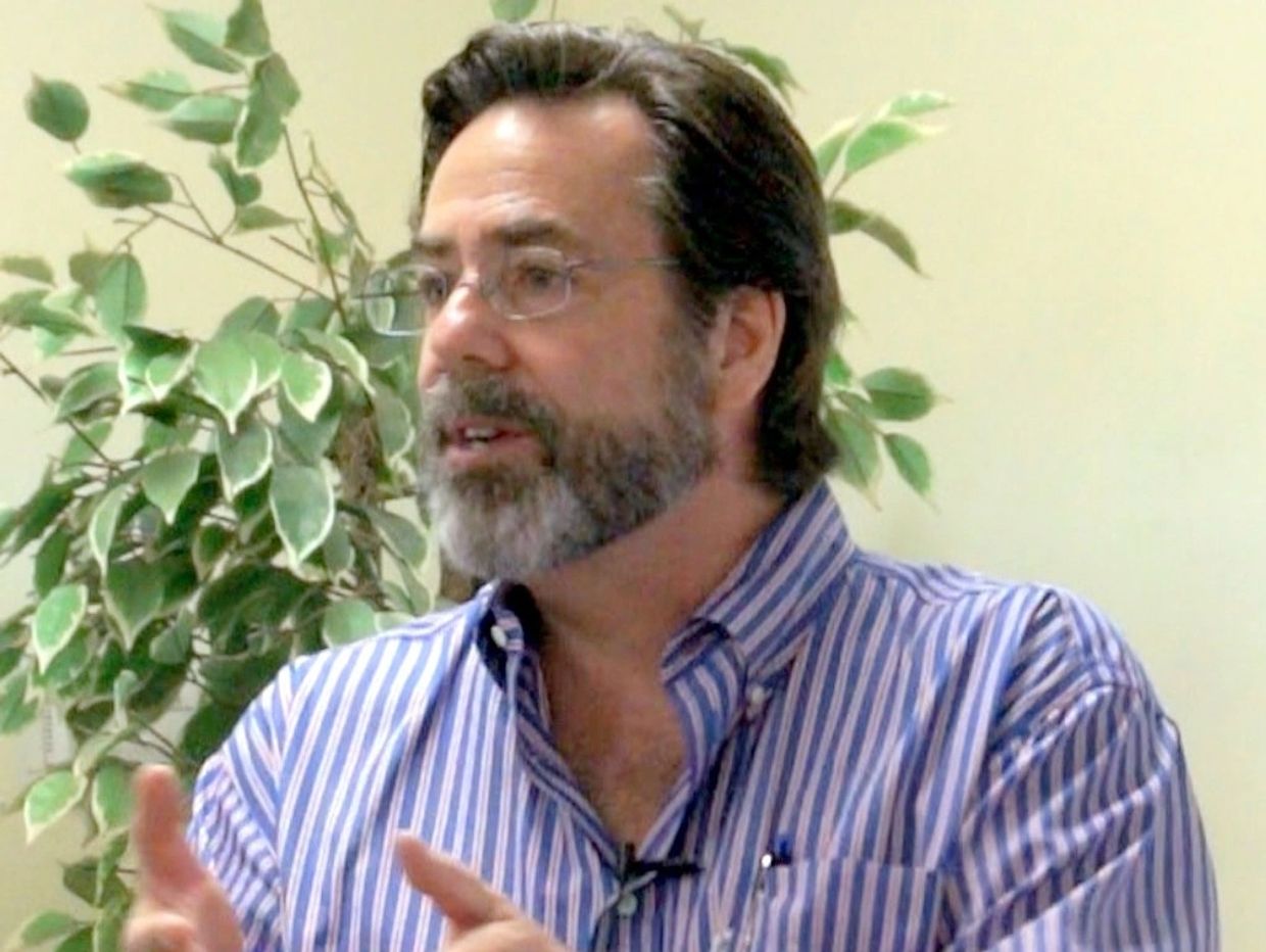 Man with glasses and beard speaking, wearing a striped shirt.