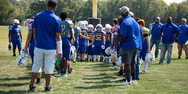 Youth football teams line up opposite each other on the field with coaches nearby.