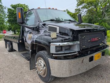 Black GMC flatbed tow truck parked on a gravel road with trees in the background.