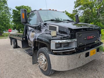 Black GMC flatbed tow truck parked on a gravel road with trees in the background.