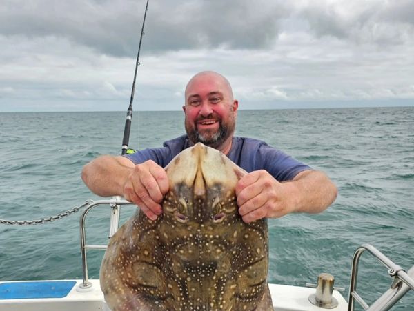 Man proudly holds a large spotted ray on a boat at sea.