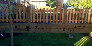 Newly installed wooden picket fence with a shovel resting against it on a sunny day.