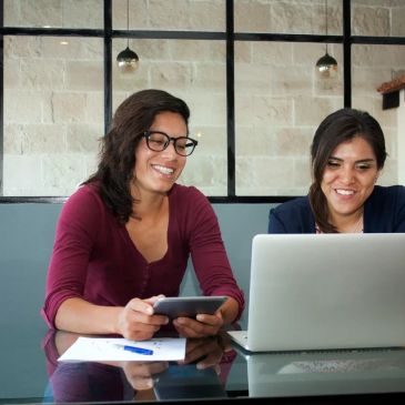 Two women smiling while using a laptop and a tablet at a desk.