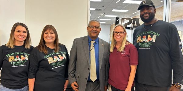 Five adults smiling indoors, some wearing 'Road to Riches Financial Literacy' shirts.