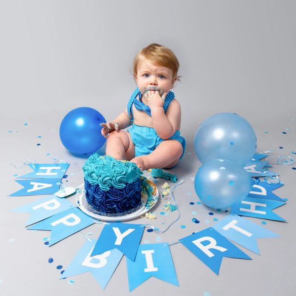 Boy dressed in a blue birthday outfit sitting eating a cake with balloons and confetti around him