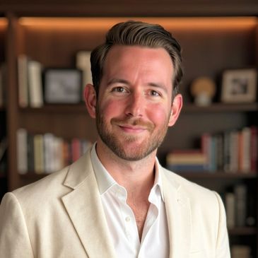 A man in a white blazer smiles in a cozy library setting.