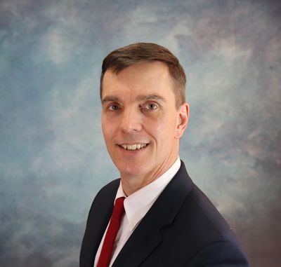 Professional headshot of a man in a suit with a red tie against a blue-gray backdrop.