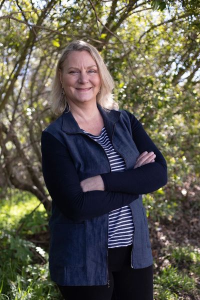 Smiling woman with blonde hair stands outdoors, arms crossed, in front of sunlit trees and greenery.