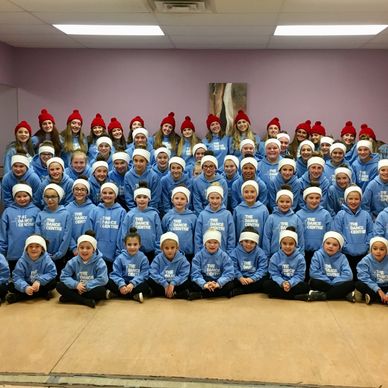Large group of children and teenagers in matching blue hoodies and red or white hats posing indoors.